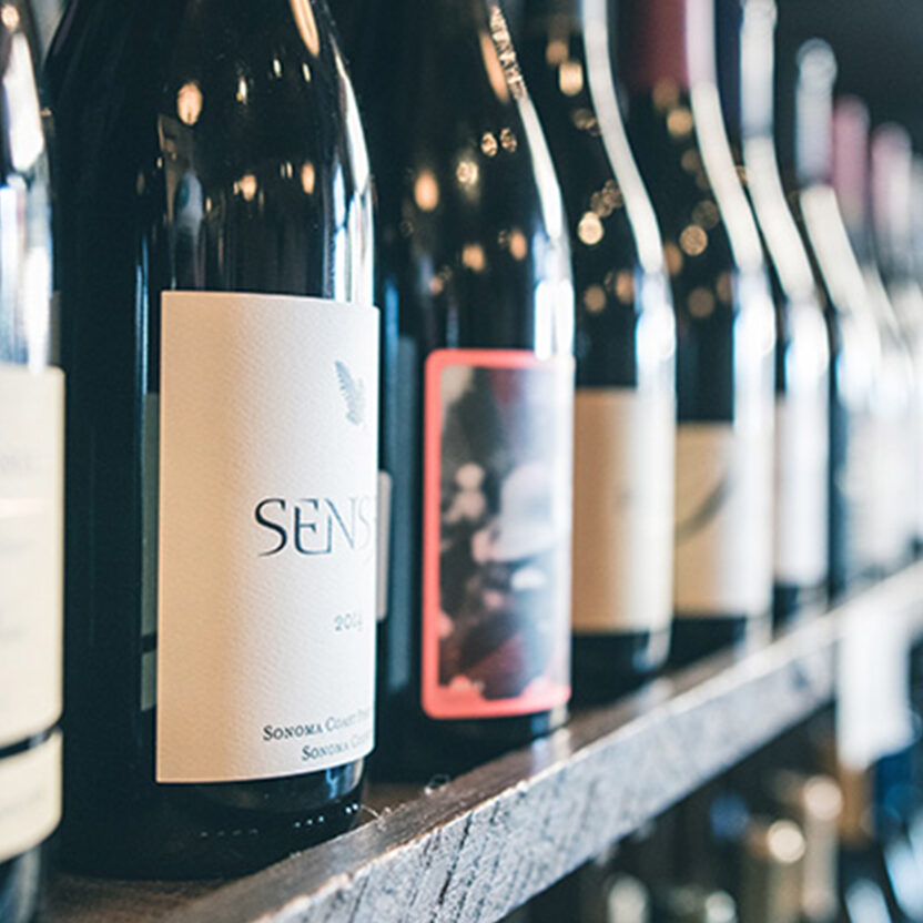 Nahaufnahme mehrerer Weinflaschen in einem Regal, Fokus auf das Etikett einer Flasche mit der Aufschrift „Sensi“.