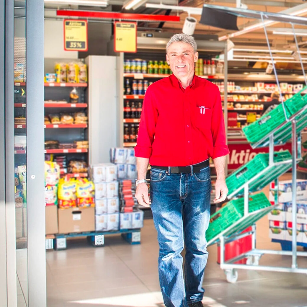Ein Mitarbeiter steht vor dem Eingang einer Denner-Filiale und lächelt in die Kamera.