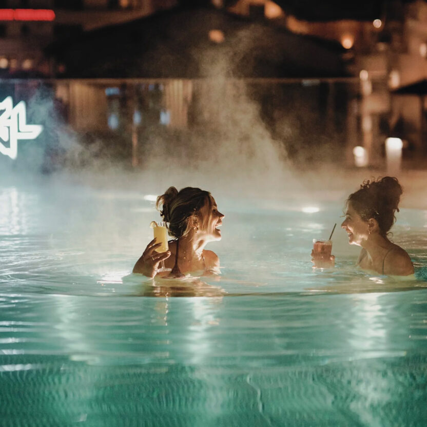 Two laughing women enjoy the warm outdoor pool of the Silvretta Therme Ischgl at night, drinks in hand and surrounded by atmospherically lit buildings.