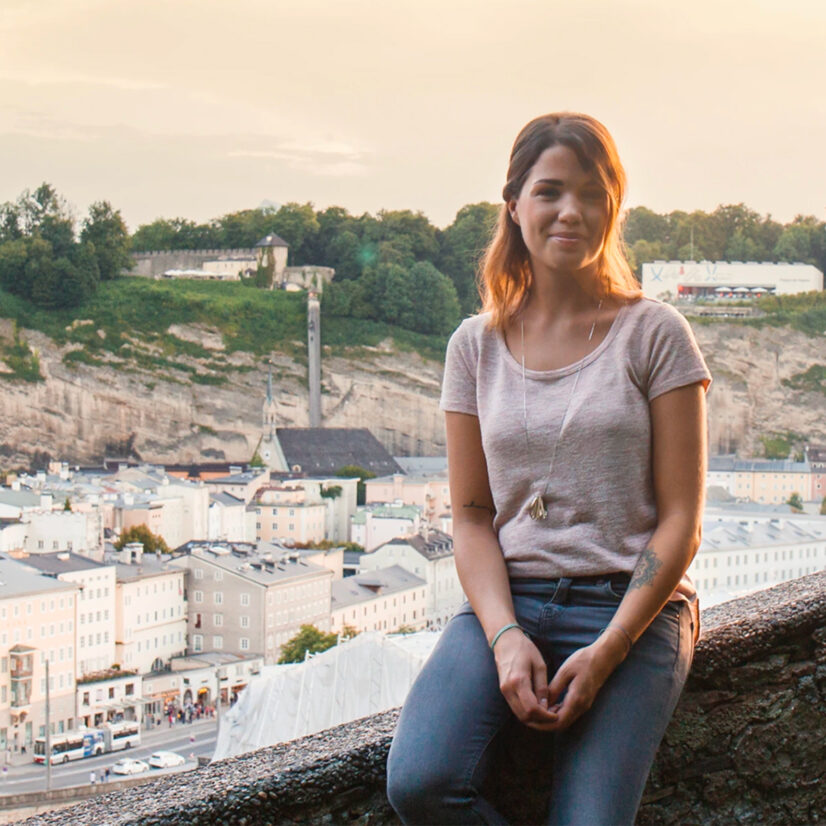Eine Frau sitzt auf einer Mauer und lächelt in die Kamera. Im Hintergrund ist die Salzburger Altstadt mit Blick auf die das Museum der Moderne.