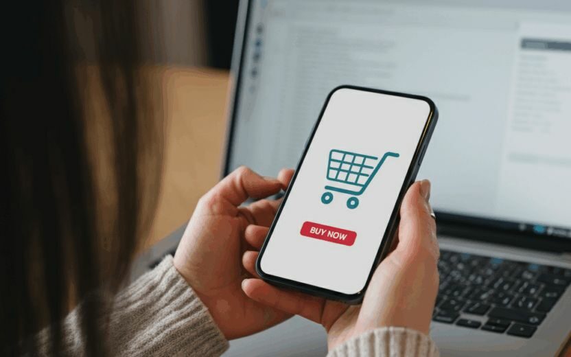 A woman holding a smartphone with the message “Buy Now” on the screen. An open laptop can be seen in the background.