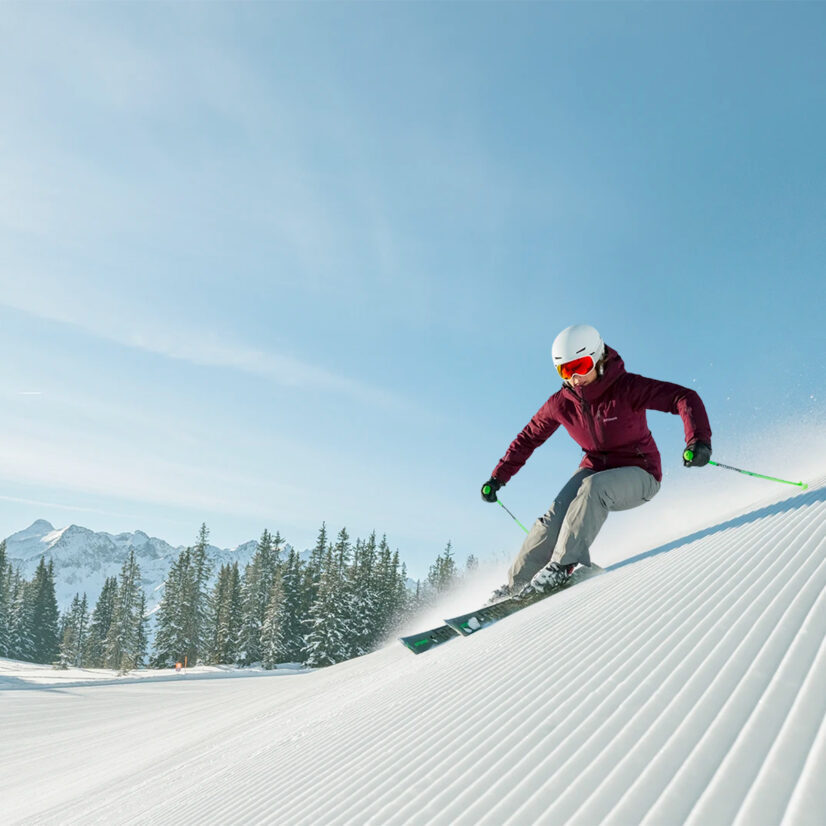 Skifahrerin rast über eine frisch präparierte Piste in Schladming Dachstein, mit verschneiten Bergen und Tannenwald im Hintergrund.