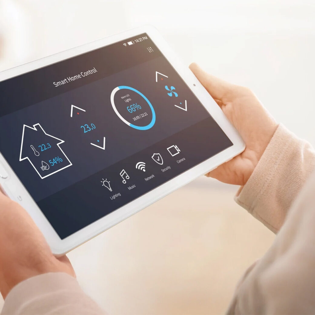 A person holds a white tablet with a smart home dashboard; displayed are temperature, a 66% circle, and icons for light, music, network, security, and energy.