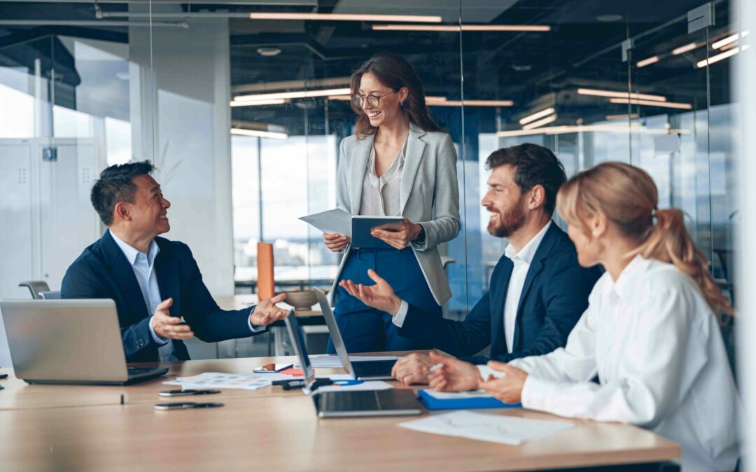 Four business people sit at a conference table with laptops and papers, while a fifth person with documents in hand leads a discussion on asset performance management (APM) in a modern office.