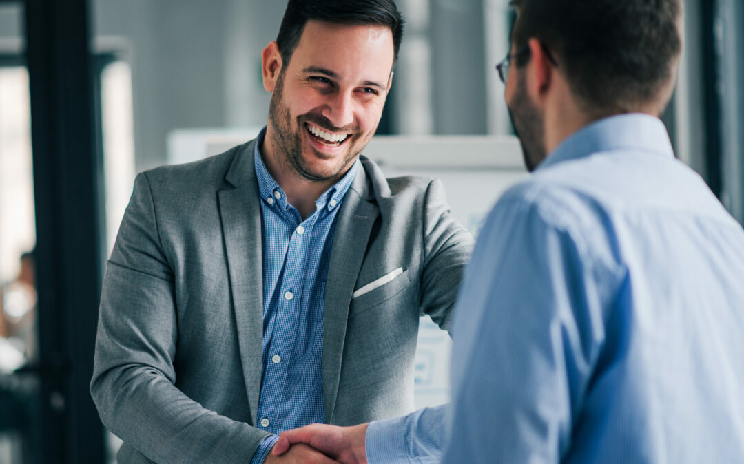 Two men in business attire are shaking hands and smiling in an office setting.