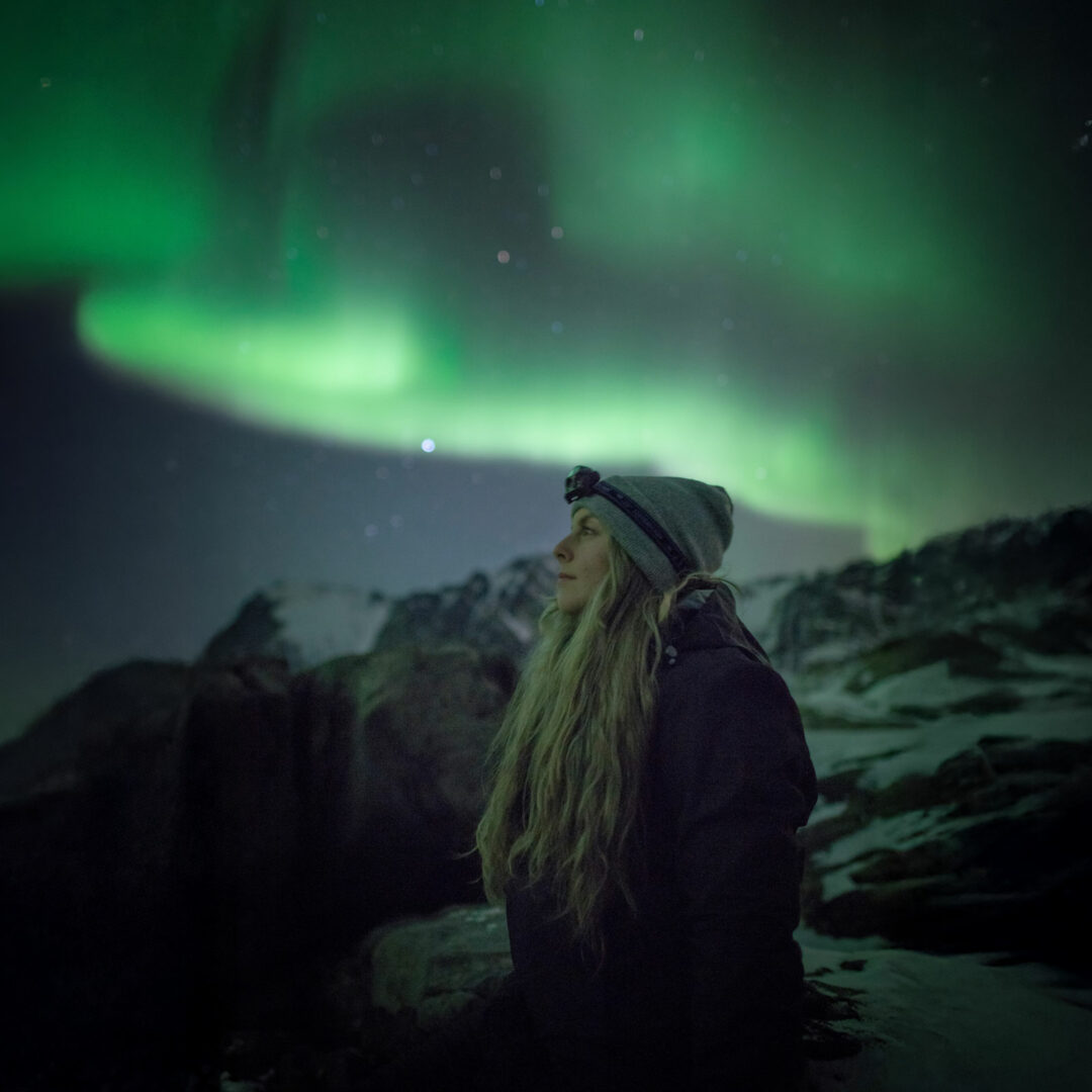 Frau steht in felsiger Umgebung und betrachtet Nordlichter