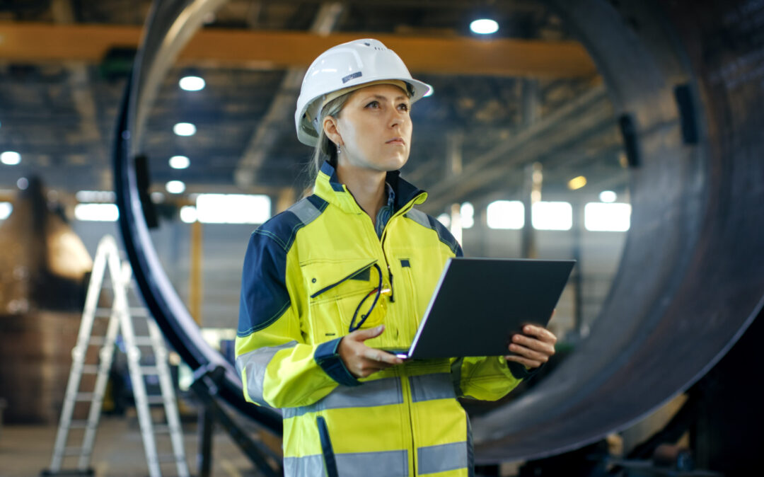 Female Industrial Engineer in the Hard Hat Uses Laptop Computer while Standing in the Heavy Industry Manufacturing Factory. In the Background Various Metalwork Project Parts Lying