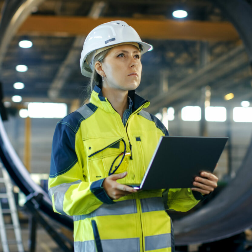 Female Industrial Engineer in the Hard Hat Uses Laptop Computer while Standing in the Heavy Industry Manufacturing Factory. In the Background Various Metalwork Project Parts Lying