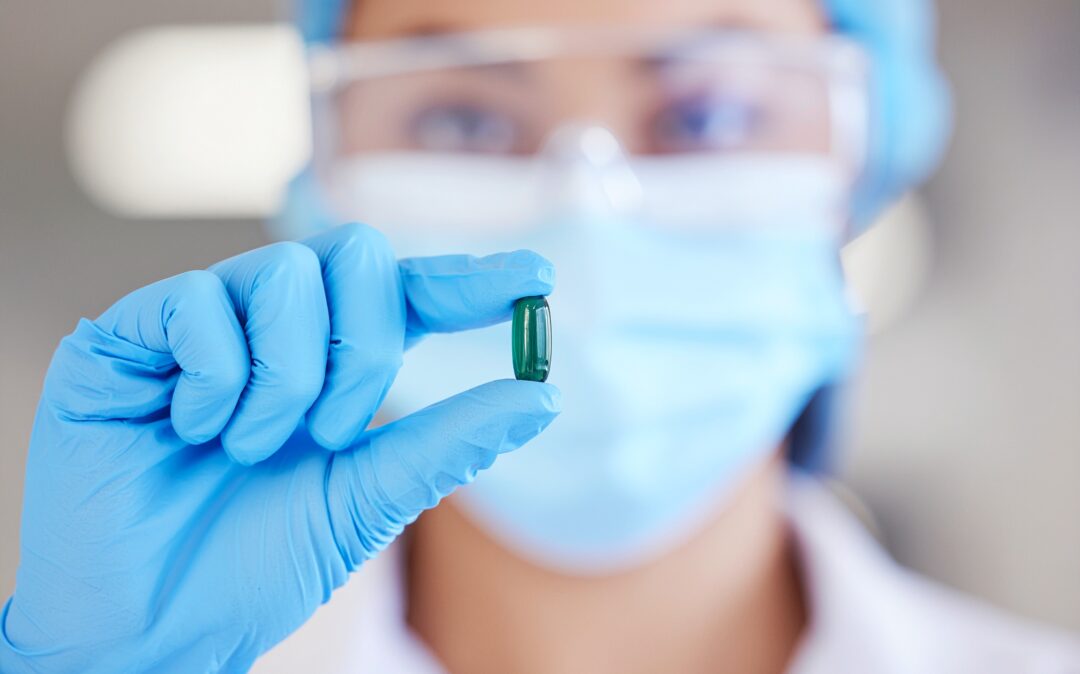 scientist wearing blue nitrile gloves and a surgical mask, holding a single translucent green capsule pill