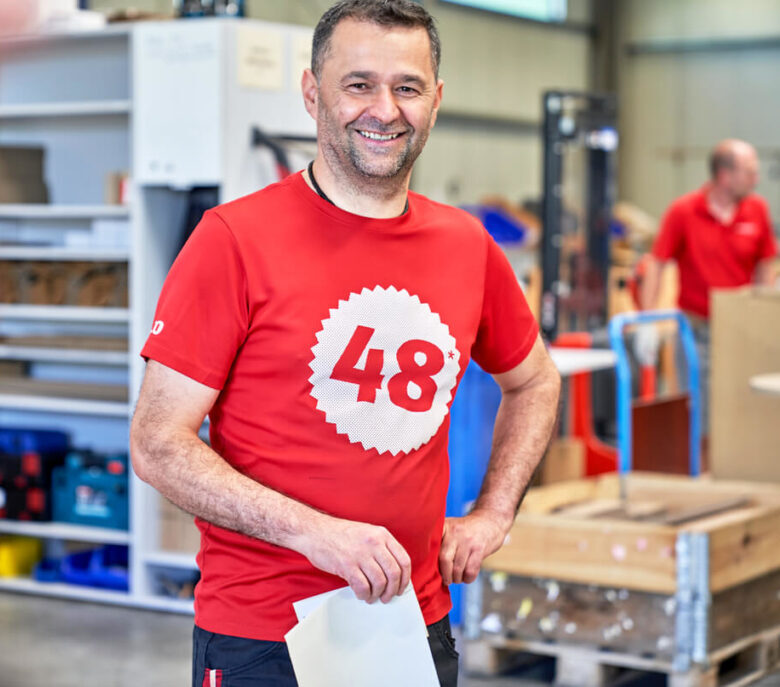 Ein Mann trägt ein rotes T-Shirt und steht in einem Lagerhaus, repräsentiert das Hammerbachers-Team.