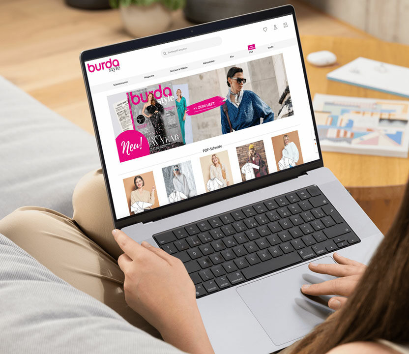 Home page of burda style on a notebook. A woman is sitting on the couch, camera looking over her shoulder. The burda style website can be seen on the screen on her lap.