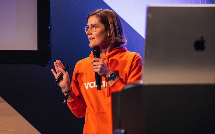 A person wearing glasses and an orange hoodie holds a microphone and gesticulates while speaking at an indoor event. A large screen and part of a monitor can be seen in the background.