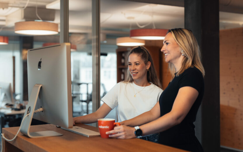 Zwei Frauen stehen an einem Stehpult und betrachten gemeinsam einen großen Bildschirm in einem modernen Büro