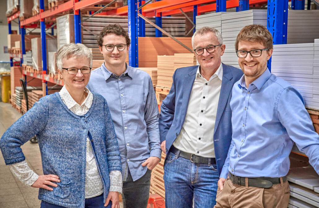Four people, three men and a woman, are standing in a warehouse with shelves in the background.