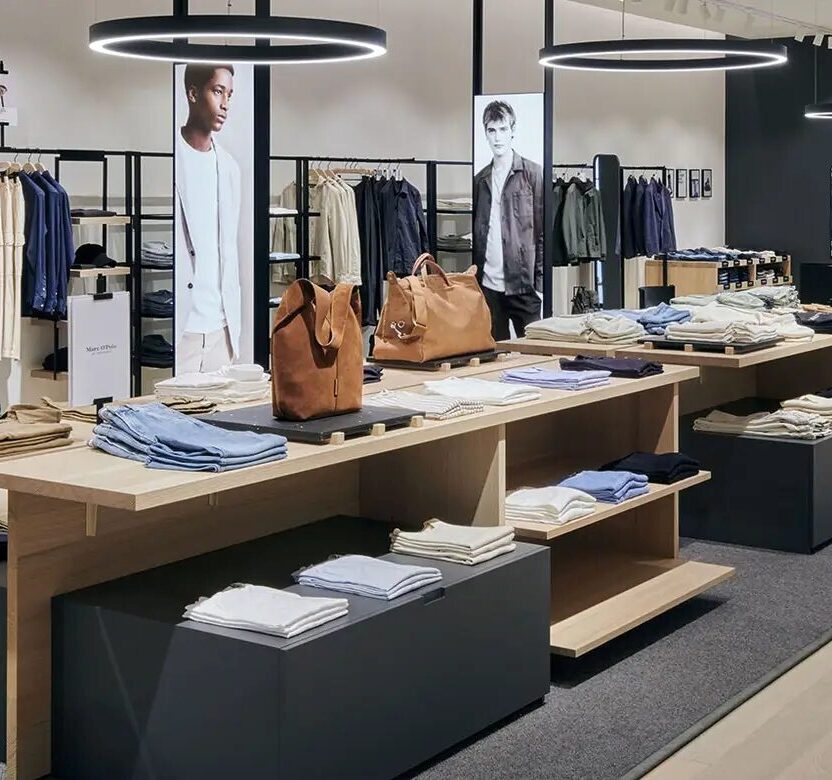 Eine Weitwinkelaufnahme eines hell beleuchteten, minimalistischen Marc O'Polo Bekleidungsgeschäfts mit Verkaufstischen aus hellem Holz, ordentlich gefalteter Kleidung und strukturierten Kleiderstangen für Herrenmode unter runden Pendelleuchten.