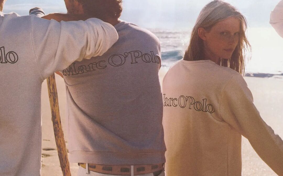 An group of four people wearing light-colored, long-sleeved shirts with the "Marc O'Polo" logo on the back, standing on a beach at sunset.