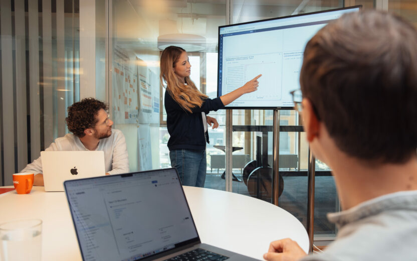 Frau zeigt auf Bildschirm mit Diagrammen und Text in einem Besprechungsraum, zwei Männer sitzen am Tisch mit geöffneten Laptops