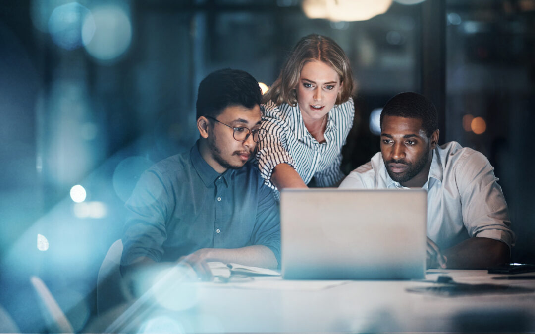Drei Personen arbeiten konzentriert zusammen an einem Laptop. Zwei Männer sitzen vor dem Bildschirm, eine Frau beugt sich zwischen ihnen nach vorne und zeigt auf den Monitor.