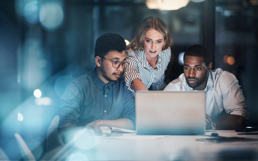 Drei Personen arbeiten konzentriert zusammen an einem Laptop. Zwei Männer sitzen vor dem Bildschirm, eine Frau beugt sich zwischen ihnen nach vorne und zeigt auf den Monitor.