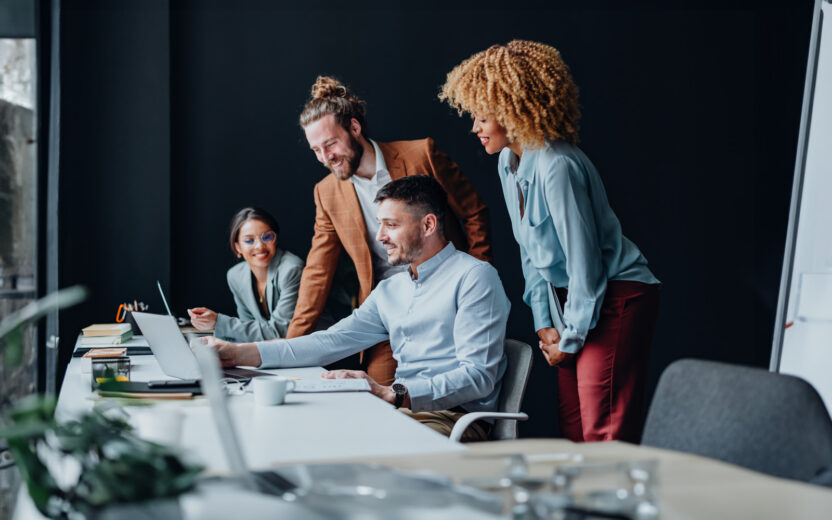 Four people are working together at a desk in an office. One man points to the screen of a laptop with an outstretched arm. His colleagues stand around him, smiling and looking at the screen