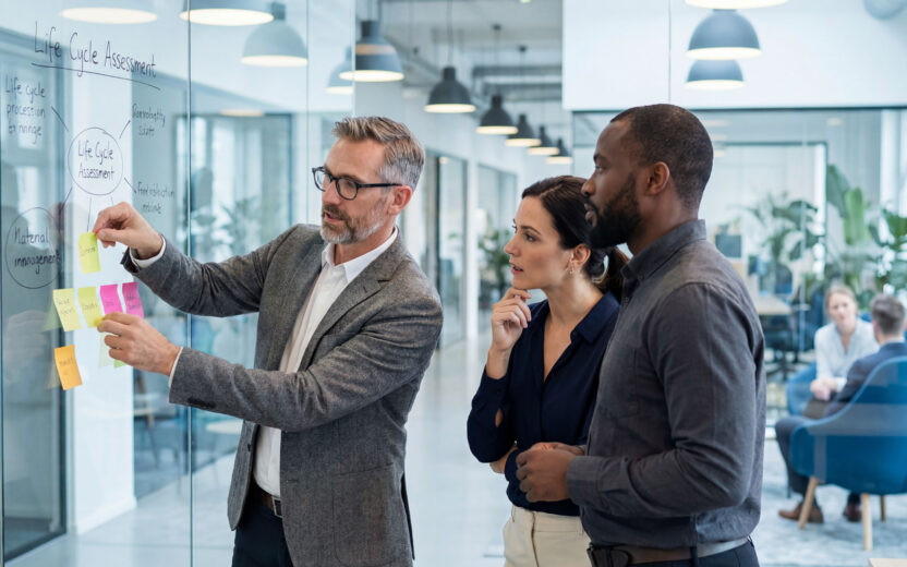 Drei Personen Drei Fachkräfte diskutieren in einem modernen Büro vor einer Glaswand mit Haftnotizen und einem Diagramm zum Thema Life Cycle Assessment (AI generated)