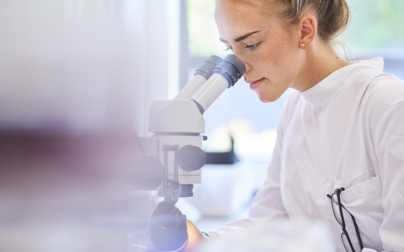 a female research scientist is analysing a sample on her microscope in a microbiology lab . the lab is brightly lit with natural light . Blurred glassware at side of frame provides copy space .