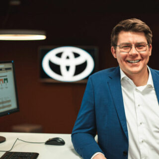 Bild von Peter-Pascal Meik, Referent auf dem Conversational AI Day am Schreibtisch lächelnd gelehnt, im Hintergrund das Logo von Toyota