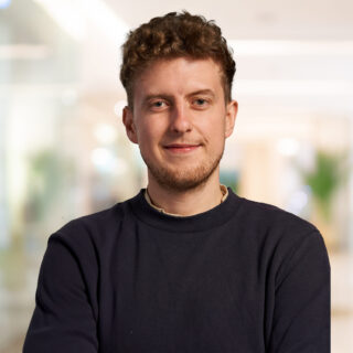Bild Johua Joel Haas, Joshua Joel Haas, Referent auf dem Conversational AI Day von valantic, lächelnder, blonder Mann mit einem dunklen Pullover