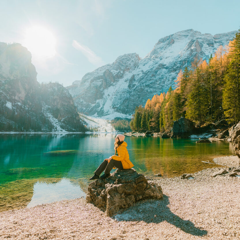 A person in a yellow jacket sits on a rock by a clear lake, surrounded by snow-capped mountains and autumn trees, under a bright sun. An AI-driven content moment that captures the serene beauty of nature.