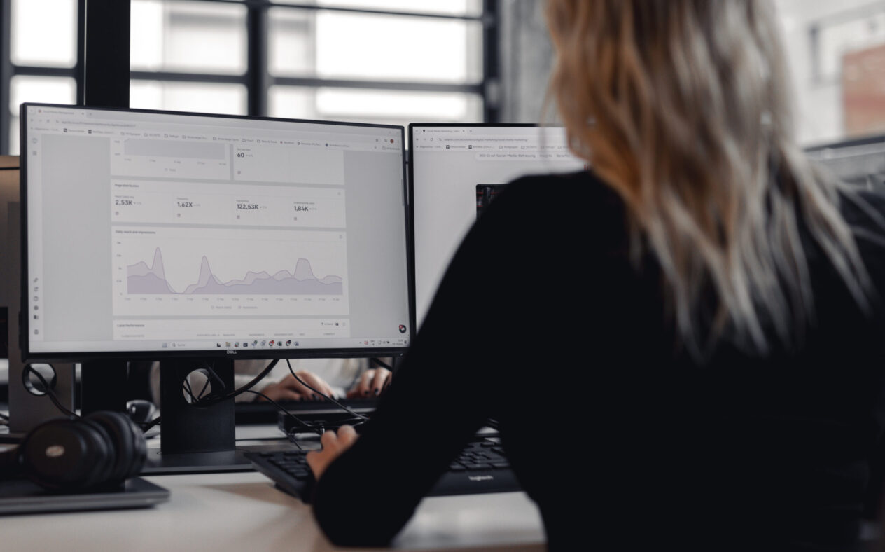 Rückansicht einer Person, die in einem modernen Büro an zwei Bildschirmen arbeitet; auf einem Monitor sind Diagramme und Analysedaten zu sehen.