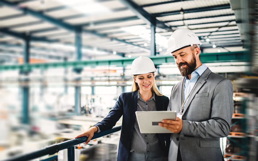 Woman and man standing on production site wearing safety helmets looking at mobile device