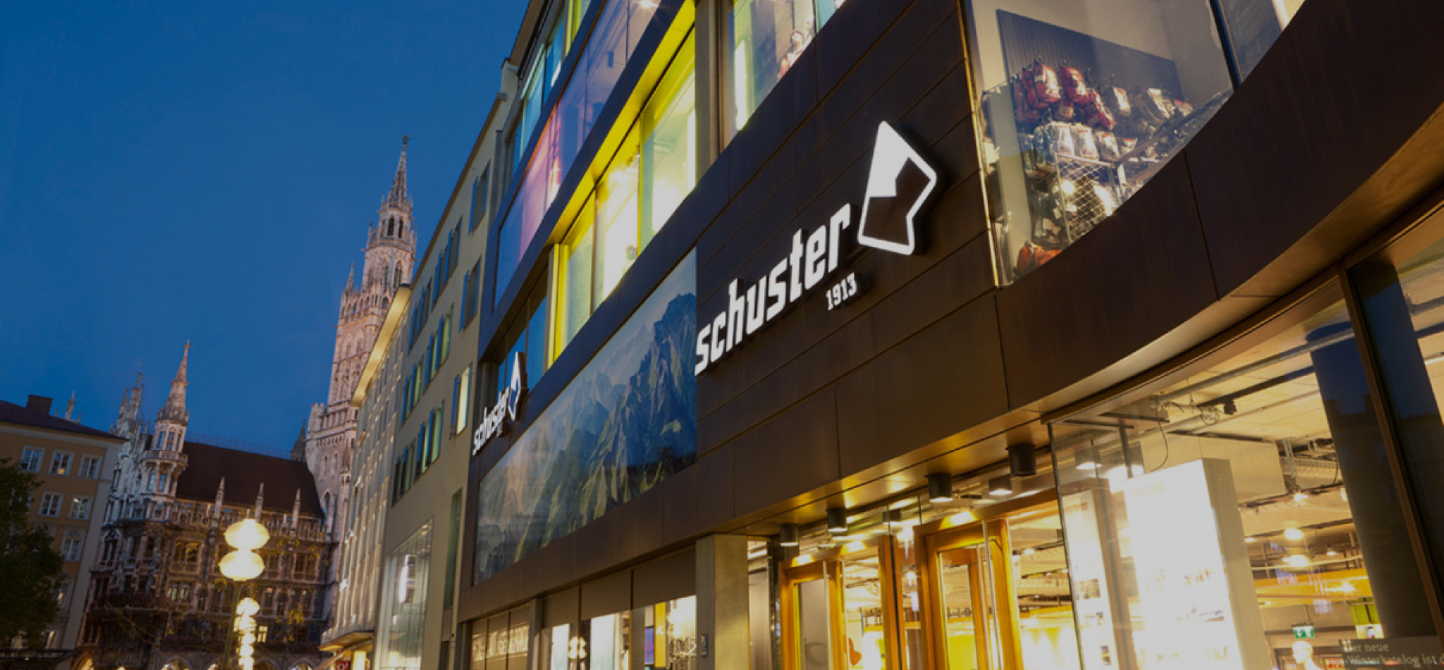 An evening exterior view of Sporthaus Schuster in Munich, featuring its illuminated facade, logo, and the Frauenkirche in the background.