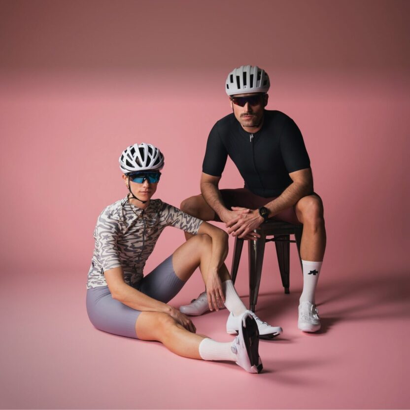 Two cyclists wearing helmets, sports glasses, and modern road cycling gear are posing in a pink studio. One person is sitting on the floor, the other on a stool. Both are wearing white cycling shoes and functional performance clothing.