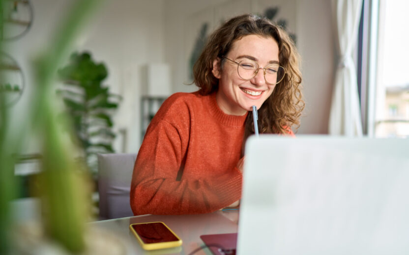 A young, happy female student using a laptop to watch a webinar while writing at home.