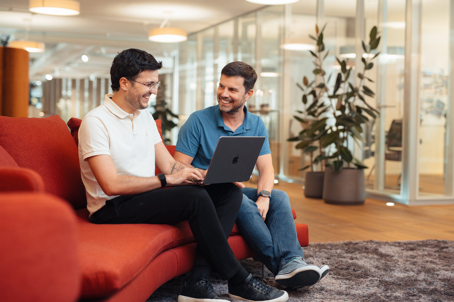 Two male professionals are sitting on a red sofa in a contemporary office setting, smiling as they look at a laptop together. In the background, you can see glass partitions, potted plants, hardwood floors, and warm overhead lighting. One man is wearing a beige polo shirt and glasses; the other is wearing a blue polo shirt. They appear to be engaged in a friendly, collaborative discussion.