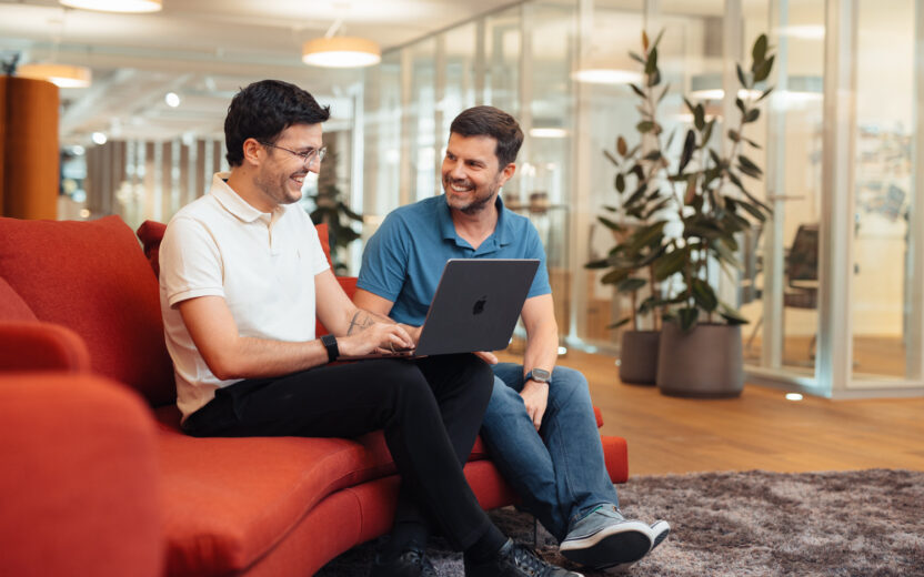 Two male professionals are sitting on a red sofa in a contemporary office setting, smiling as they look at a laptop together. In the background, you can see glass partitions, potted plants, hardwood floors, and warm overhead lighting. One man is wearing a beige polo shirt and glasses; the other is wearing a blue polo shirt. They appear to be engaged in a friendly, collaborative discussion.