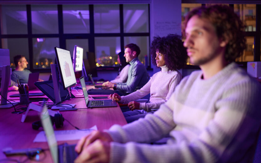 Colleagues work at computer monitors and laptops in a purple-lit modern office at night