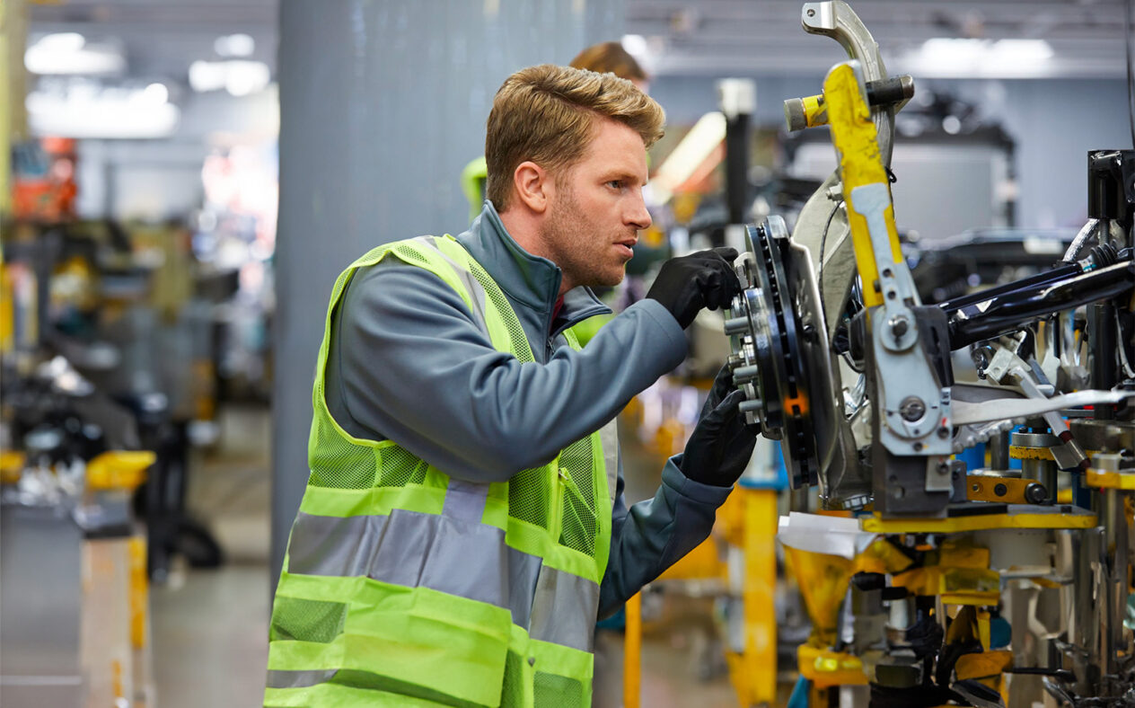 Ein Mechaniker mit neongelber Warnweste und Schutzhandschuhen inspiziert konzentriert die Radaufhängung und Bremsscheibe eines Fahrzeugs in einer Automobilfertigungshalle.