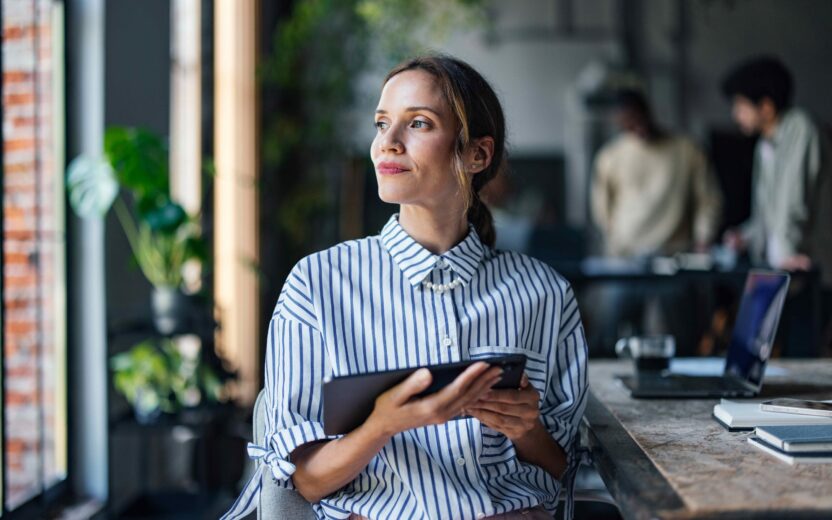 Young woman holding a tablet and coffee cup in a modern café, looking thoughtfully out the window