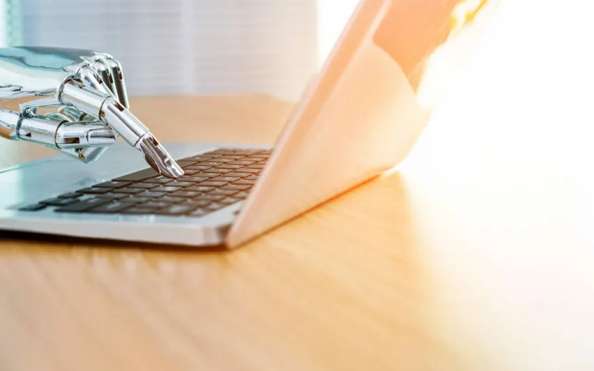 Eine Roboterhand tippt auf einer Laptoptastatur auf einem Holzschreibtisch, im Hintergrund strömt Sonnenlicht. Sie verkörpert die nahtlose Integration von Technologie und Natur. Diese Szene unterstreicht die Möglichkeiten der Konversations-KI bei der Transformation alltäglicher Aufgaben.