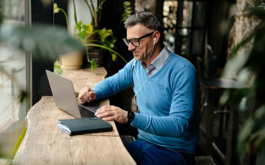 Adult smiling man in glasses and headphones working with laptop while sitting in cafe