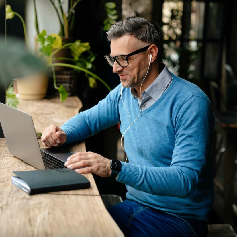 Adult smiling man in glasses and headphones working with laptop while sitting in cafe