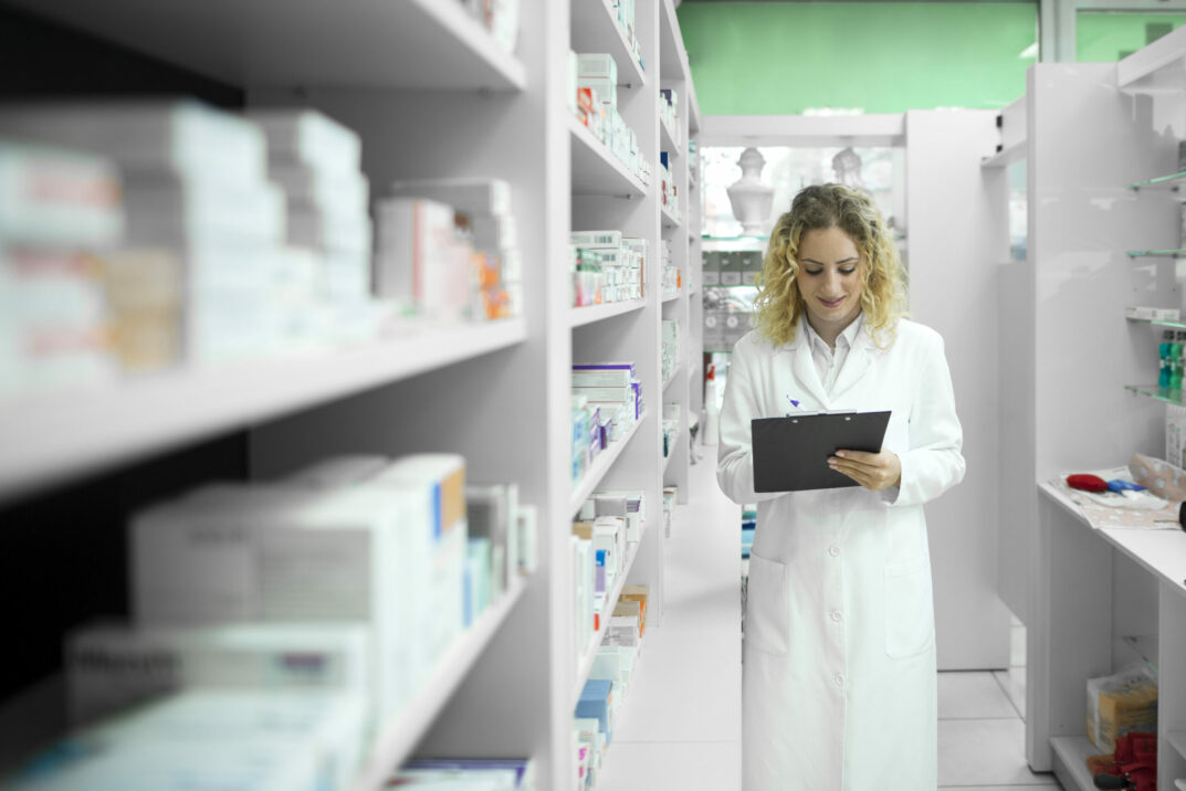 Frau steht mit weißem Kittel in einem Raum mit Medikamenten in den Regalen und notiert etwas auf einem Klemmbrett