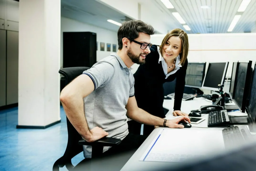Image of a man sitting at a desk and a smiling woman looking at him
