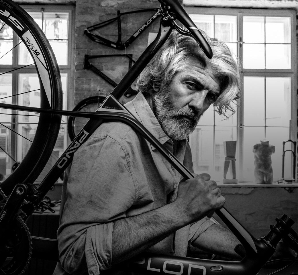 Picture of an elderly gentleman in a bicycle repair shop shouldering a SIMPON bicycle.