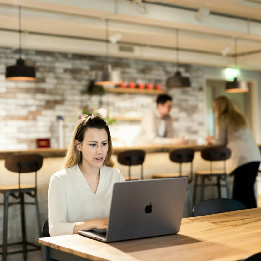 Frau, die an einem Holztisch am Laptop arbeitet - im Hintergrund ist eine Bar