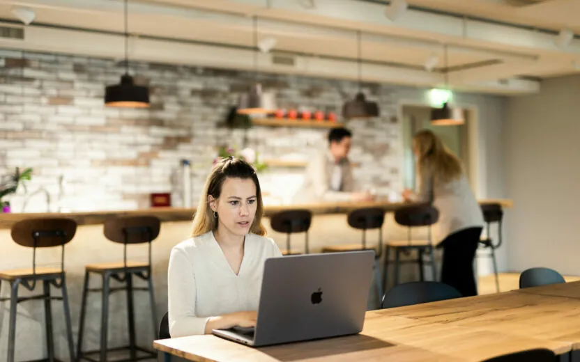 Frau, die an einem Holztisch am Laptop arbeitet - im Hintergrund ist eine Bar
