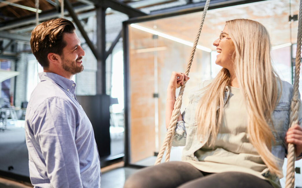 Picture of a laughing woman on an office swing and a man standing next to her.
