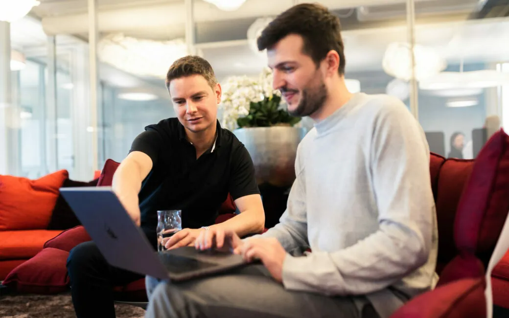 Zwei Männer auf gepolsterten Sesseln sitzen und zusammen an einem Laptop arbeiten.
