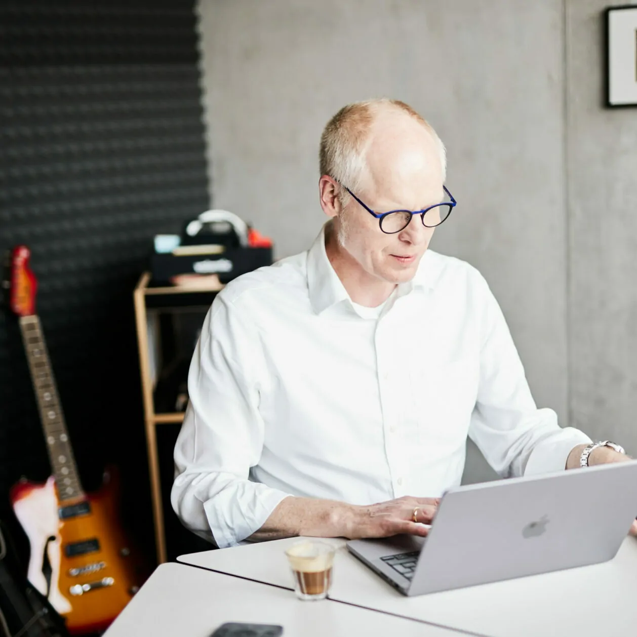 Ein älterer Mann, der am Laptop arbeitet und nach seinem Smartphone greift - im Hintergrund sind E-Gitarren zu sehen.